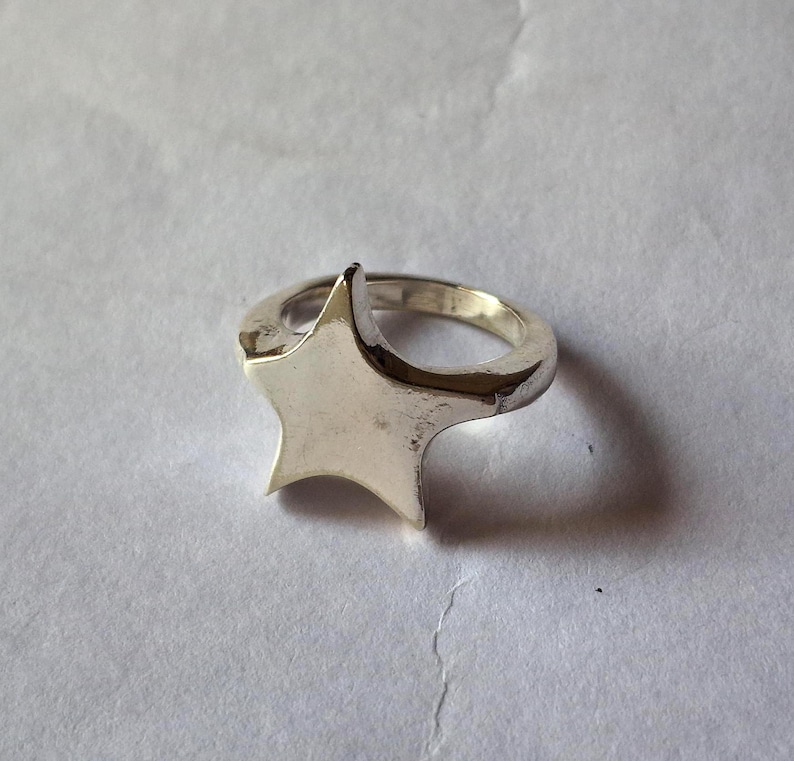 Op de afbeelding: Een zilverkleurige ring met een sterontwerp. De ster is het middelpunt, met een glad, gepolijst oppervlak. De ringband is ook zilver en heeft een eenvoudig, afgerond ontwerp. De ring staat tegen een neutrale achtergrond.
