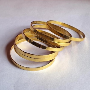 May include: A collection of six gold-colored bangles, each with a simple, rounded design. The bangles are stacked and slightly overlapping, creating a layered effect. The jewelry is set against a plain white background.