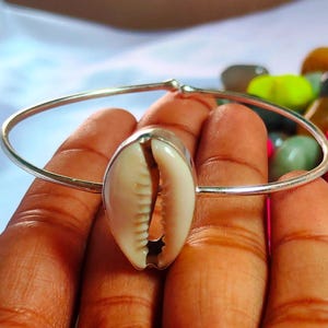 Può includere: Un braccialetto a cerchio argentato con un elemento centrale a forma di conchiglia naturale color crema. Il braccialetto è tenuto in una mano, con pietre colorate sullo sfondo. La conchiglia ha una superficie strutturata e un interno scuro.