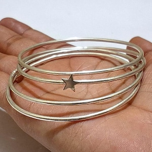 Peut inclure: Ensemble de cinq bracelets argentés fins et ronds, dont un orné d'une petite étoile. Les bracelets sont empilés sur une main. Bijoux simples et élégants, adaptés au quotidien.