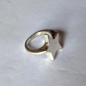 Op de afbeelding: Een zilverkleurige ring met een stervormig ontwerp. De ster is verhoogd en heeft vijf punten. De ringband is glad en cirkelvormig. De ring is waarschijnlijk gemaakt van metaal en staat tegen een witte achtergrond.