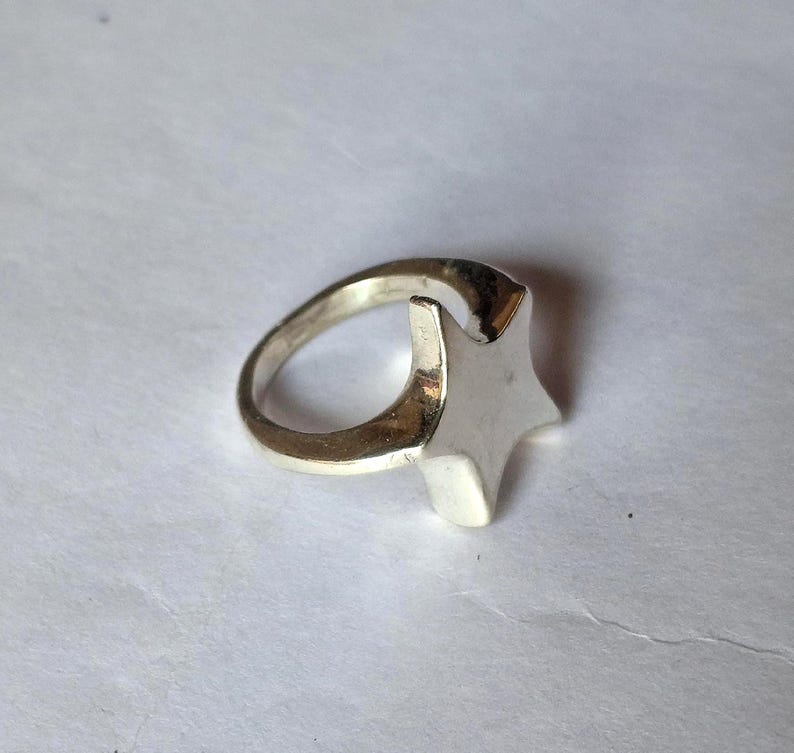 Op de afbeelding: Een zilverkleurige ring met een stervormig ontwerp. De ster is verhoogd en heeft vijf punten. De ringband is glad en cirkelvormig. De ring is waarschijnlijk gemaakt van metaal en staat tegen een witte achtergrond.
