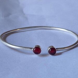 Könnte beinhalten: Ein silberfarbenes offenes Armreif-Armband mit einem roten Edelstein an jedem Ende. Das Armband hat ein schlichtes, elegantes Design mit glatter, polierter Oberfläche. Die Edelsteine sind rund und tiefrot.