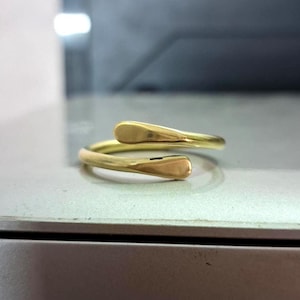 Puede incluir: Un anillo dorado con un diseño abierto único. El anillo presenta dos extremos redondeados y aplanados que se curvan suavemente alejándose el uno del otro. El anillo es de metal brillante y reflectante.