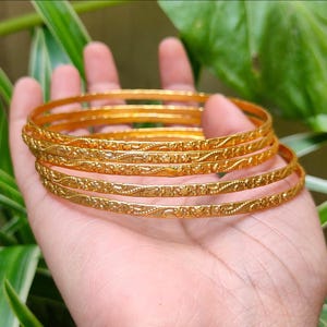 Könnte beinhalten: Ein Satz von sechs goldenen Armreifen mit aufwendigen floralen Mustern. Die Armreifen sind auf einer Hand gestapelt und zeigen ihr detailliertes Design und ihre glänzende Oberfläche. Der Hintergrund zeigt grünes Laub.
