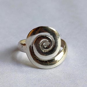 May include: A silver ring with a spiral design. The ring features a polished, swirling pattern in the centre, resembling a nautilus shell. The band and the spiral are made of silver. The ring is set against a white background.