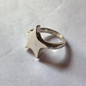 Op de afbeelding: Een zilverkleurige ring met een stervormig ontwerp. De ring heeft een gepolijste afwerking en een eenvoudige band. De ster heeft puntige randen en een plat oppervlak. De ring staat tegen een witte achtergrond.