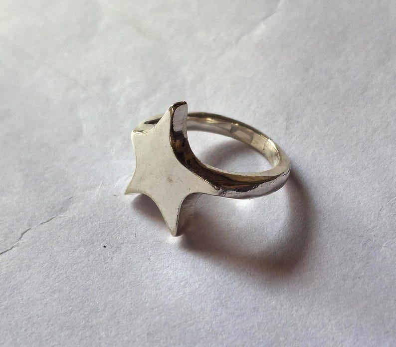 Op de afbeelding: Een zilverkleurige ring met een stervormig ontwerp. De ring heeft een gepolijste afwerking en een eenvoudige band. De ster heeft puntige randen en een plat oppervlak. De ring staat tegen een witte achtergrond.