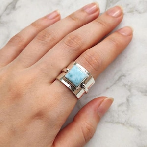 May include: A wide sterling silver ring featuring a square, light blue, marbled stone set in the center. The band has a polished, reflective finish and a smooth, thick construction that wraps around the finger with a prominent metal setting.
