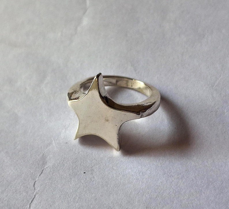 Op de afbeelding: Een zilverkleurige ring met een vijfpunts sterontwerp. De ster is het middelpunt, met een glad, gepolijst oppervlak. De ringband is ook zilverkleurig, wat een samenhangende uitstraling cre&euml;ert.
