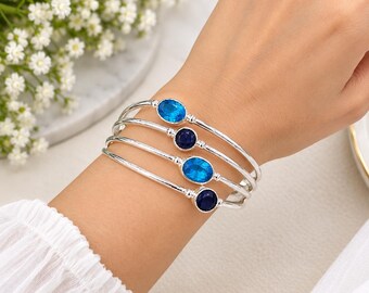 May include: A set of four silver-toned bangles, each featuring a single oval-shaped gemstone. Two bangles are set with bright blue stones, and two bangles are set with dark navy blue stones. The bracelets have a smooth, polished metallic finish.