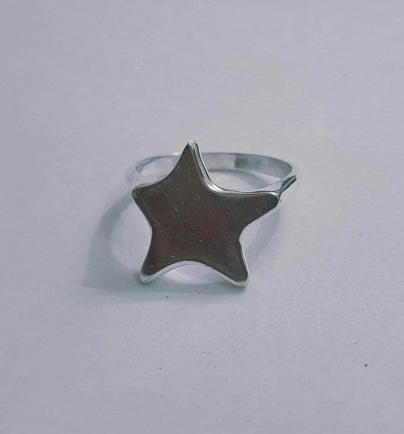 Op de afbeelding: Een zilverkleurige ring met een stervormig ontwerp. De ster is het middelpunt, met een glad, metalen oppervlak. De ringband is eenvoudig en past bij de vorm van de ster. Dit sieraad is geschikt voor dagelijks gebruik.