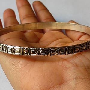 Puede incluir: Un brazalete plateado con un patrón geométrico repetido alrededor de la banda. El brazalete circular parece ser de metal. El brazalete se sostiene en una mano y el fondo es de color neutro.