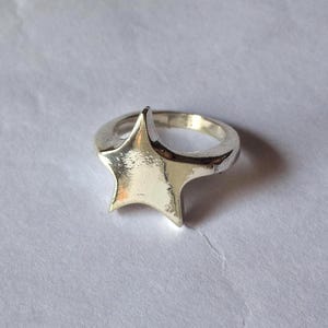 Op de afbeelding: Een zilveren ring met een sterontwerp. De ring heeft een dikke band en een driedimensionale ster. De ster is glad en glanzend, met een licht getextureerd oppervlak. De ring staat tegen een witte achtergrond.