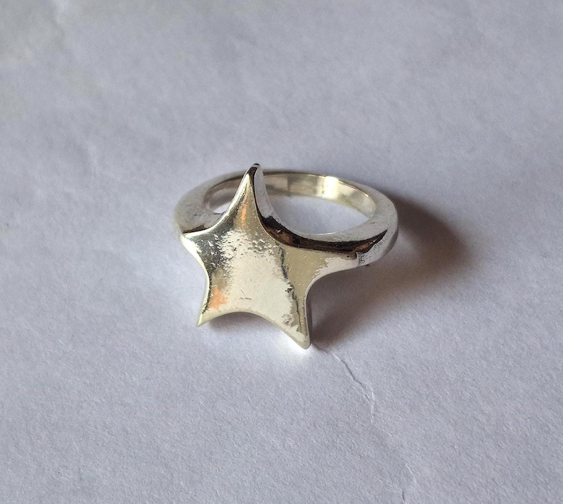 Op de afbeelding: Een zilveren ring met een sterontwerp. De ring heeft een dikke band en een driedimensionale ster. De ster is glad en glanzend, met een licht getextureerd oppervlak. De ring staat tegen een witte achtergrond.