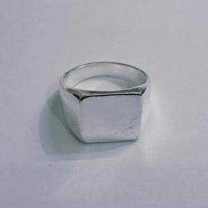 May include: A silver signet ring with a square face. The ring has a thick band and a polished, reflective surface. A classic piece of jewellery.