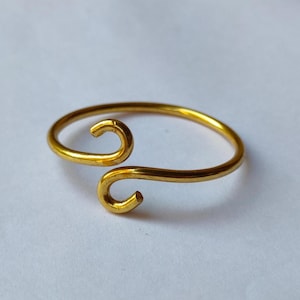 May include: A gold-colored ring with a unique, open design. The ring features two curved, hook-like ends that meet in the middle. The ring is made of a shiny, metallic material and is set against a white background.