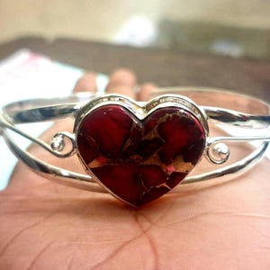 Puede incluir: Brazalete de plata con una piedra roja y dorada en forma de corazón. El corazón es el punto focal, engastado en un marco plateado con adornos en espiral. El brazalete se sostiene en una mano, mostrando su diseño.
