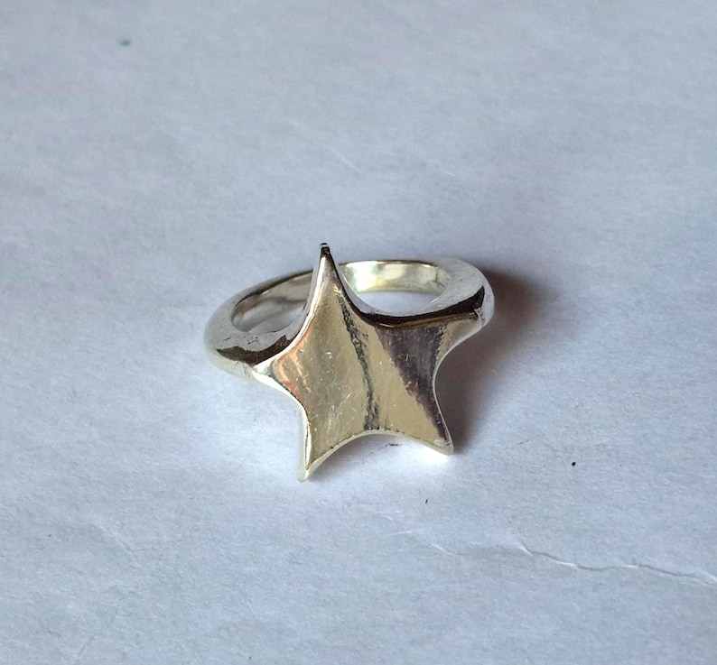 Op de afbeelding: Een zilverkleurige ring met een stervormig ontwerp. De ster heeft vijf punten en is het middelpunt van de ring. De ringband is glad en afgerond, en de ster is verhoogd.