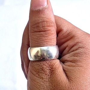 May include: A wide, polished silver ring is worn on a thumb. The ring has a smooth, rounded surface that reflects light. The ring is a simple, classic design, suitable for everyday wear. The ring is a piece of jewellery.