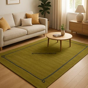 May include: A rectangular olive green rug with a dark blue border and corner detailing. The rug is placed in a living room setting with a beige sofa, wooden coffee table, and a window with white curtains. The text "BESPOKECUSTOMRUGS" is visible on the rug.