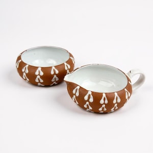 May include: Two ceramic serving pieces. One is a small bowl, the other a creamer with a handle and spout. Both are brown with a white teardrop pattern and white interiors. The pieces are on a white background.