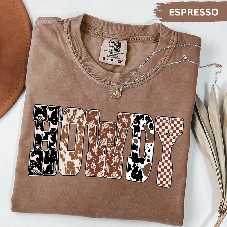 K&ouml;nnte beinhalten: Espressofarbenes Comfort Colors T-Shirt mit dem Wort "HOWDY" in einem Western-Design. Die Buchstaben zeigen Muster wie Kuhdruck, Kakteen und ein Schachbrettmuster. Eine silberne Halskette mit Anh&auml;nger ist &uuml;ber dem Shirt drapiert.