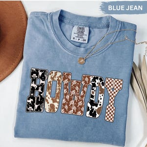 K&ouml;nnte beinhalten: Ein blaues Comfort Colors T-Shirt mit dem Wort "HOWDY" in einem Western-Design. Die Buchstaben zeigen Kuhmuster, Kakteen und Schachbrettmuster. Eine goldene Halskette mit Anh&auml;nger liegt auf dem Shirt.