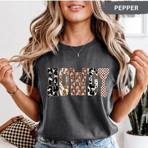 K&ouml;nnte beinhalten: Dunkelgraues T-Shirt mit dem Wort "HOWDY" in einem Western-Design. Die Buchstaben zeigen Kuhmuster, Kakteen und Karomuster. Das Shirt ist ein l&auml;ssiger Stil.