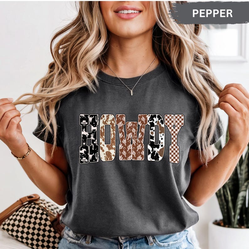 K&ouml;nnte beinhalten: Dunkelgraues T-Shirt mit dem Wort "HOWDY" in einem Western-Design. Die Buchstaben zeigen Kuhmuster, Kakteen und Karomuster. Das Shirt ist ein l&auml;ssiger Stil.