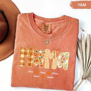 Könnte beinhalten: Ein korallenfarbenes Comfort Colors T-Shirt mit dem Wort "Mama" in einer Mischung aus Karomustern und Blumenmustern. Die Namen Liam, Noah, Olivia, Emma und Amelia sind darunter mit Kürbis-Grafiken aufgedruckt.