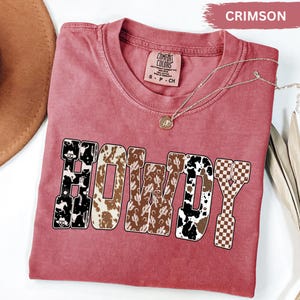 K&ouml;nnte beinhalten: Karmesinrotes T-Shirt mit dem Wort "HOWDY" in einem Western-Design. Die Buchstaben zeigen Muster wie Kuhfell, Kakteen und ein Schachbrettmuster. Eine goldene Halskette mit Anh&auml;nger ist &uuml;ber dem Shirt drapiert.