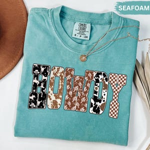K&ouml;nnte beinhalten: T&uuml;rkisfarbenes T-Shirt mit dem Wort "HOWDY" in einem Western-Design. Die Buchstaben weisen Muster wie Kuhfell, Kakteen und ein Schachbrettmuster auf. Eine goldene Halskette mit Anh&auml;nger ist auf dem T-Shirt drapiert.