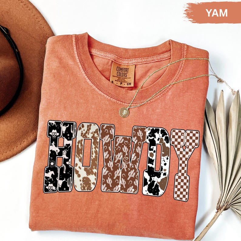 K&ouml;nnte beinhalten: Ein orangefarbenes Comfort Colors T-Shirt mit dem Wort "HOWDY" in einem Western-Design. Die Buchstaben zeigen Muster wie Kuhdruck, Kakteen und ein Schachbrettmuster. Eine goldene Halskette und ein brauner Hut sind ebenfalls sichtbar.