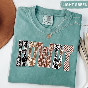 K&ouml;nnte beinhalten: Ein hellgr&uuml;nes Comfort Colors T-Shirt mit dem Wort "HOWDY" in einer kr&auml;ftigen, dekorativen Schriftart. Die Buchstaben weisen verschiedene Muster auf, darunter Kuhfell, Kakteen und ein kariertes Design. Eine silberne Halskette mit Anh&auml;nger liegt auf dem Shirt.