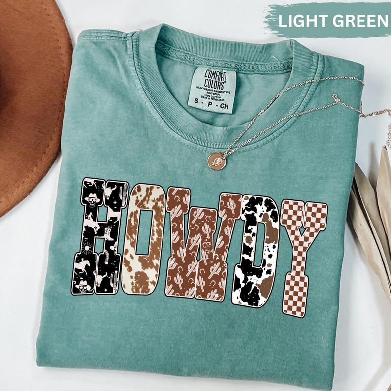 K&ouml;nnte beinhalten: Ein hellgr&uuml;nes Comfort Colors T-Shirt mit dem Wort "HOWDY" in einer kr&auml;ftigen, dekorativen Schriftart. Die Buchstaben weisen verschiedene Muster auf, darunter Kuhfell, Kakteen und ein kariertes Design. Eine silberne Halskette mit Anh&auml;nger liegt auf dem Shirt.