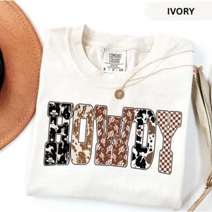 K&ouml;nnte beinhalten: Elfenbeinfarbenes T-Shirt mit dem Wort "HOWDY" in einer dekorativen Schriftart. Die Buchstaben weisen verschiedene Muster auf, darunter Kuhfellmuster, Kakteen und ein Schachbrettmuster. Eine goldene Halskette mit Anh&auml;nger liegt auf dem Shirt.