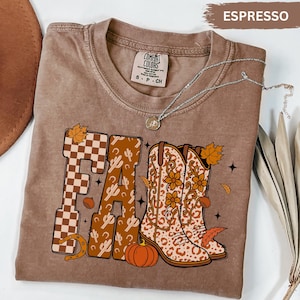 May include: Espresso-coloured t-shirt with a "Fall" graphic featuring a checkered "F", cactus-patterned "A" and "L", and cowboy boots with floral and leopard print. Includes a pumpkin, horseshoe, and leaves.