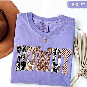 K&ouml;nnte beinhalten: Ein violettes Comfort Colors T-Shirt mit dem Wort "HOWDY" in einem Western-Design. Die Buchstaben zeigen Muster wie Kuhfell, Kakteen und ein Schachbrettmuster. Eine goldene Halskette mit Anh&auml;nger ist sichtbar.