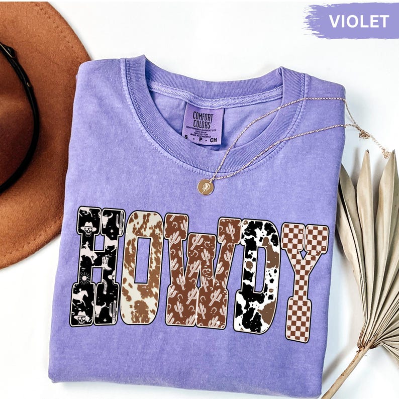 K&ouml;nnte beinhalten: Ein violettes Comfort Colors T-Shirt mit dem Wort "HOWDY" in einem Western-Design. Die Buchstaben zeigen Muster wie Kuhfell, Kakteen und ein Schachbrettmuster. Eine goldene Halskette mit Anh&auml;nger ist sichtbar.