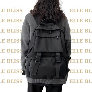 May include: A black backpack featuring a large main compartment, a smaller zippered front pocket, and two vertical straps with plastic buckle closures. The bag has a top handle and adjustable shoulder straps, presented in a solid black color.