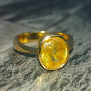 May include: A gold ring featuring a large, oval-cut yellow gemstone. The ring has a simple, polished band and a bezel setting. The bright yellow stone is set against a textured surface.
