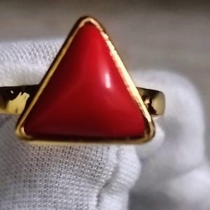 May include: A gold ring featuring a vibrant red triangular stone. The stone is set in a gold band, creating a striking contrast. The ring's design is simple yet elegant, making it a versatile accessory.