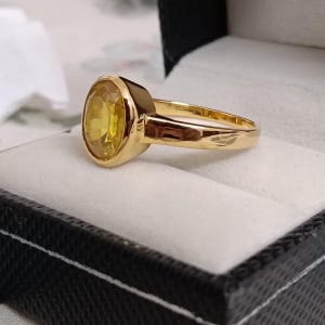 May include: A gold ring featuring a large, oval yellow gemstone. The ring is set in a polished gold band and presented in a black jewellery box with a white interior. The gemstone has a bright, clear appearance. The ring is a classic design.