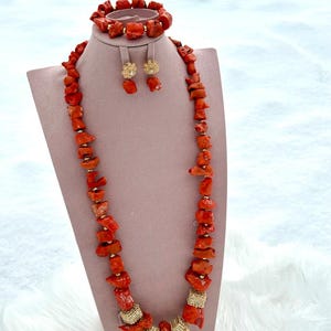 May include: A coral jewelry set displayed on a pink mannequin. The set includes a necklace, bracelet, and earrings. The necklace and bracelet are made of irregular coral pieces with gold accents. The earrings are gold with a floral design. The background is white.