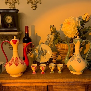 Puede incluir: Una colección de piezas de cerámica antiguas, que incluye dos jarras decorativas con asas y tapas rojas, un plato y varias tazas pequeñas. Las piezas están dispuestas sobre un mueble de madera, con un reloj y flores en el fondo.