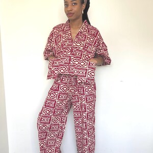 May include: A two-piece matching set consisting of a short-sleeved button-down shirt with a collar and wide-leg trousers. The garment features a repeating geometric pattern in deep red and off-white, with a structured fit and front pockets.