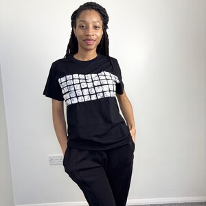 Può includere: T-shirt nero con un motivo geometrico bianco sul petto. La persona indossa pantaloni neri. Lo sfondo è un muro bianco e un tappeto grigio.