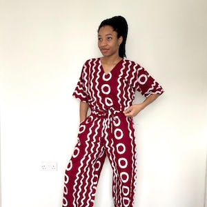 May include: A burgundy two-piece outfit with a white wavy and circle pattern. The top has short sleeves and a V-neck, and the pants are wide-legged. The outfit is tied at the waist with a matching fabric belt.