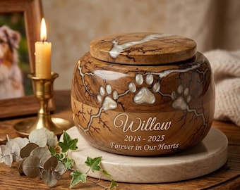May include: A wooden pet urn with a lid, featuring paw prints and the name "Willow" with dates and the phrase "Forever in Our Hearts." The urn is brown with white accents and sits on a marble base.